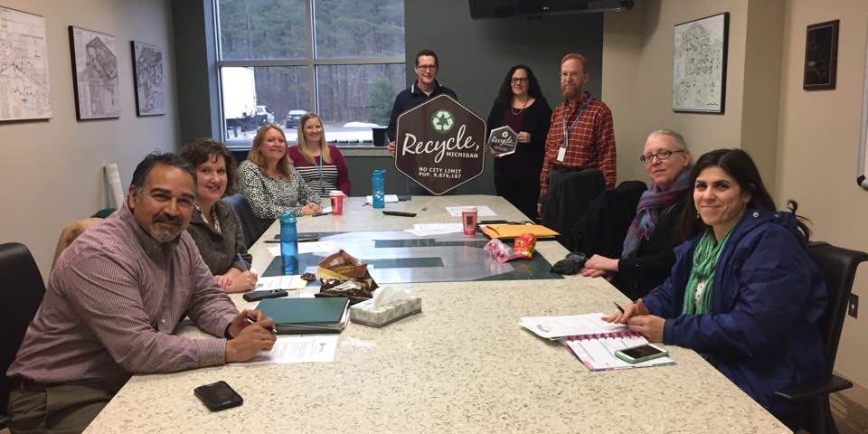 Members of the Regional Recycling Coordinating Committee sitting around a table with a Recycle, Michigan sign.